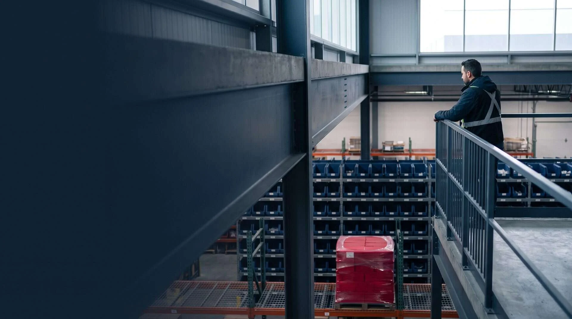 Operations manager on a warehouse mezzanine overlooking organized shelving with navy bins and a crimson-wrapped pallet