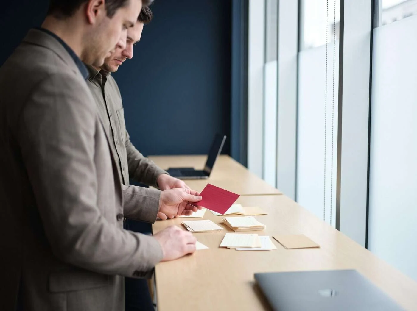 Deux collègues debout à un poste de travail près d’une fenêtre trient des fiches de mots-clés en groupes, l’un tenant une carte cramoisie