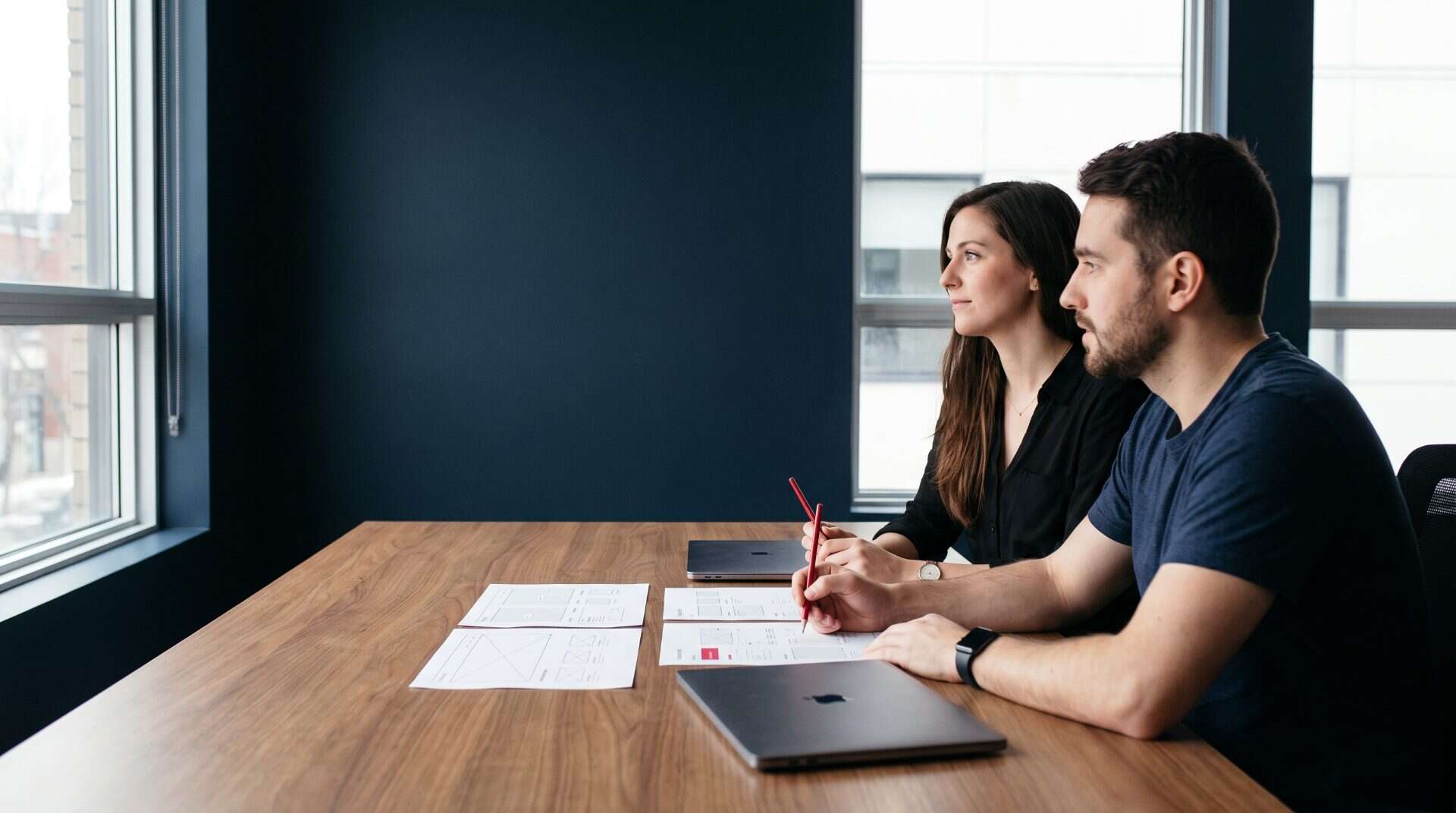 Product designer and developer side by side at a walnut table reviewing printed website wireframes, one holding a crimson pencil, navy wall behind
