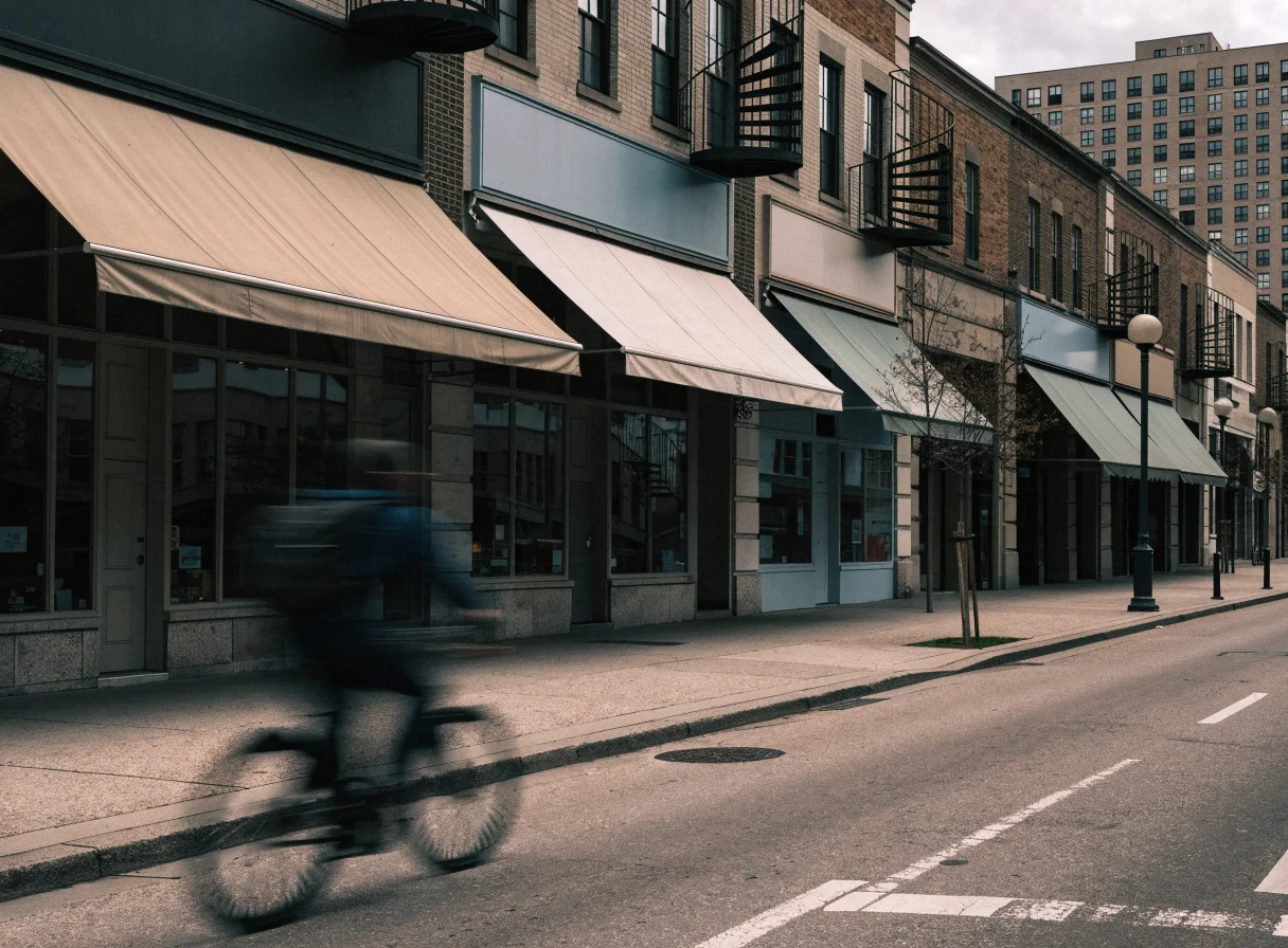 A row of independent ground-floor shops on a Montreal street, with fabric awnings, blank unlettered sign panels, wrought-iron spiral staircases above, and a cyclist passing in motion blur under overcast light.