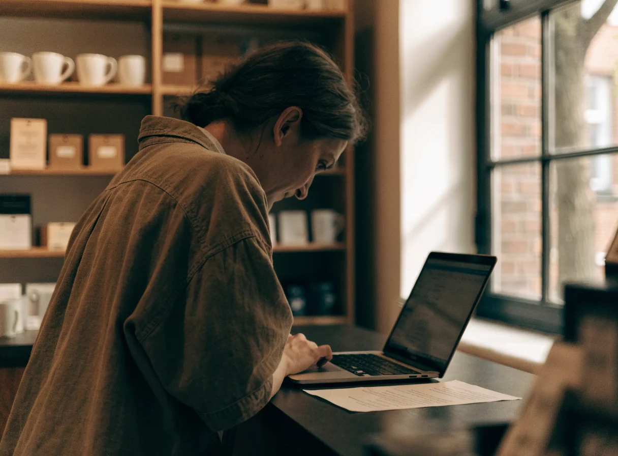A small business owner leaning over a shop counter, reading an open laptop screen with a printed document beside it, warm shelving out of focus behind and daylight from a tall window.