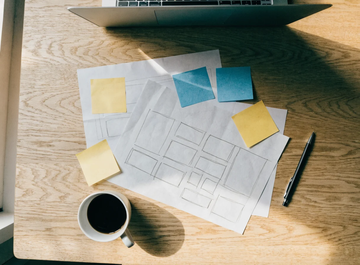 Overhead view of a website planning session: pencil wireframe sketches of page layouts on plain paper, yellow and blue sticky notes, a pen, a cup of black coffee, and the edge of an open laptop on a light oak table.